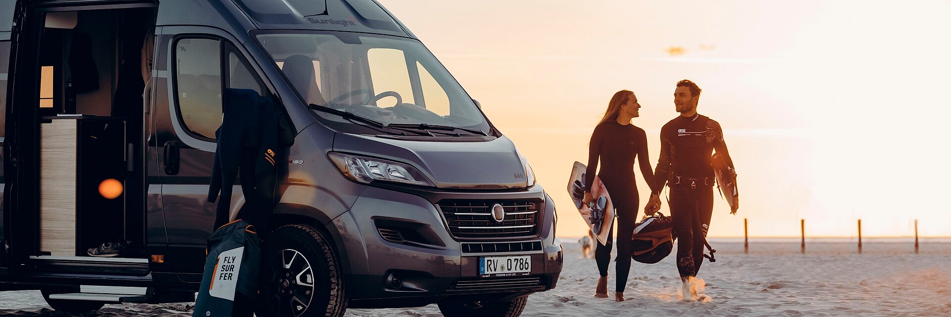 Schwarzer Sunlight Campervan geparkt auf einem weitläufigen Strandabschnitt. Davor ein Surfer-Pärchen im Sonnenuntergang.