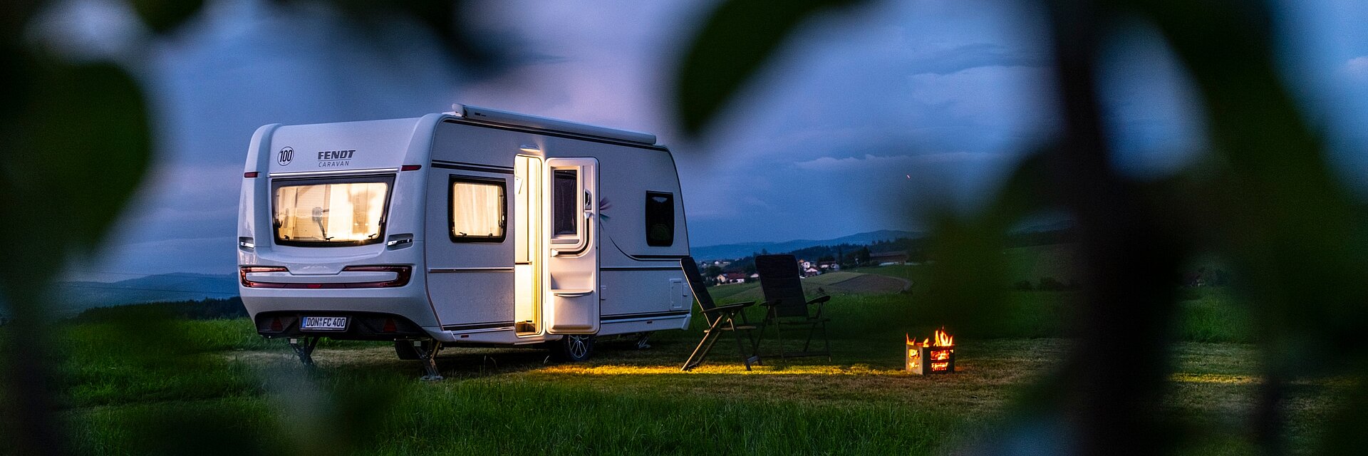 Von innen beleuchteter weißer Fendt Wohnwagen bei Dunkelheit auf einem Feld. Davor zwei Campingstühle und ein Feuer.
