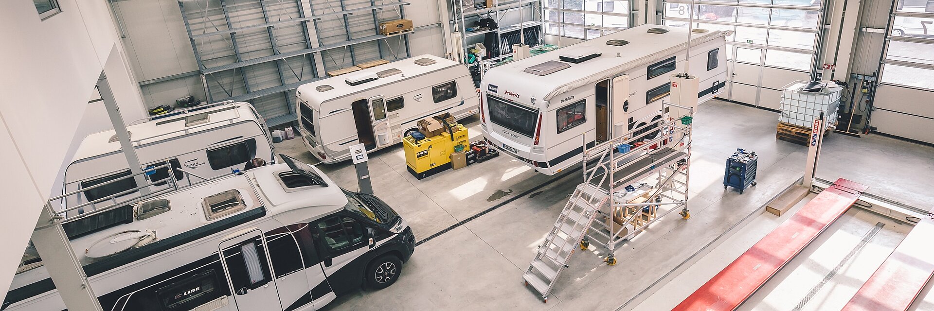 Innenaufnahme der Werkstatt von Ullrich Caravaning mit drei Wohnwagen und einem Campervan sowie einer leeren roten Hebebühne.