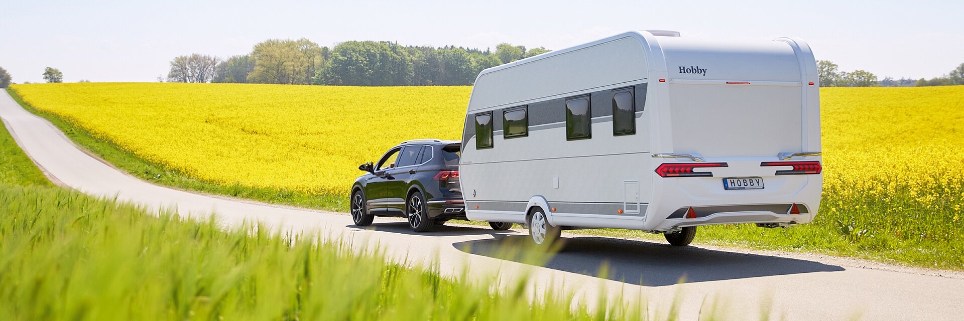 Weißer Hobby Wohnwagen, gezogen von einem schwarzen Auto vor einem gelb blühendem Rapsfeld in sonniger Kulisse.