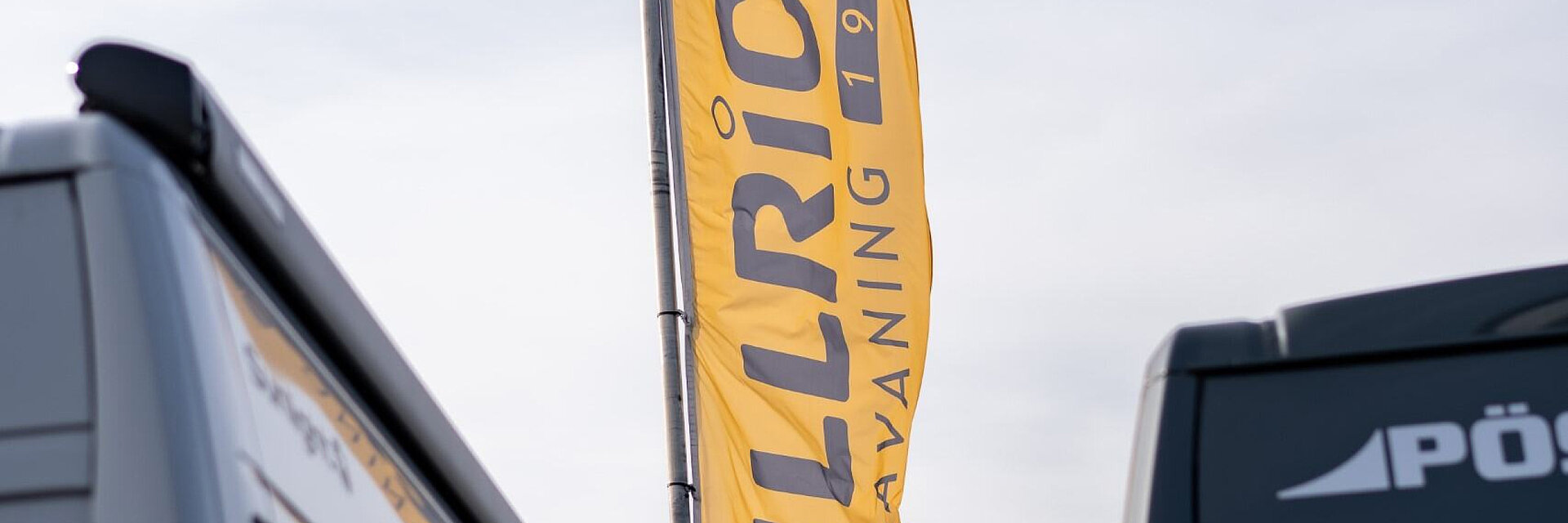 Außenaufnahme einer gelben Flagge von Ullrich Caravaning zwischen zwei Camping-Fahrzeugen vor blauem Himmel.
