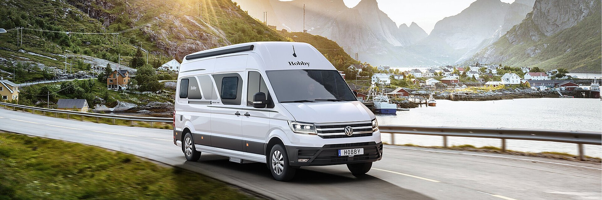 Weißer Hobby Campervan auf einer Straße vor einer Gebirgslandschaft mit angrenzendem Fischerdorf an einem See.