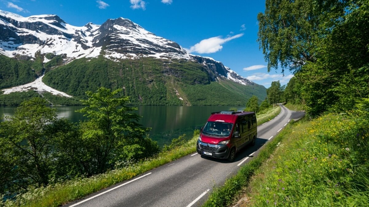 Roter Pössl Campervan fahrend auf einer sonnigen Gebirgsstraße entlang eines Sees.