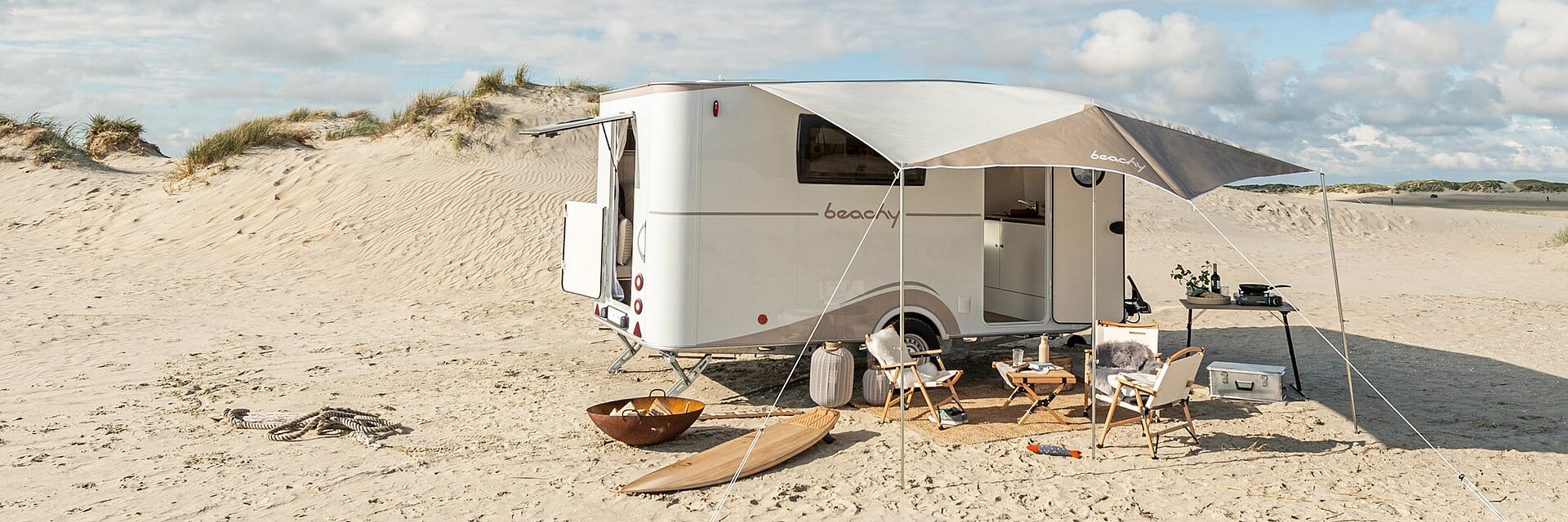 Weißer Beachy Wohnwagen mit aufgebauter Markise sowie Stühlen, Tischen und Surfbrettern auf einem sonnigen Strandabschnitt.